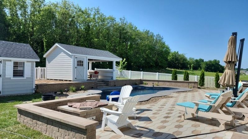 Backyard pool area with white sheds, patio, and chairs under an umbrella. Green trees in background.