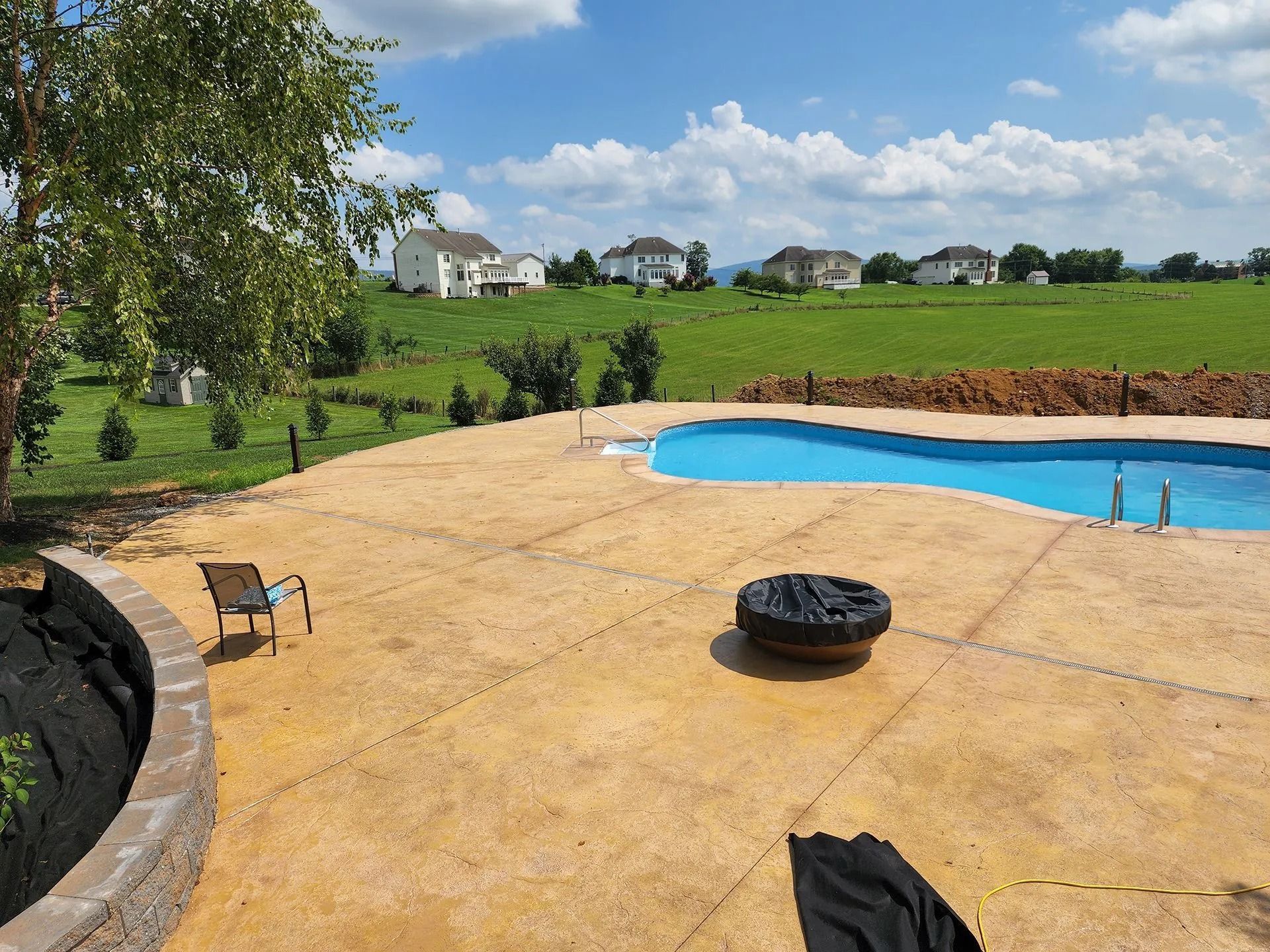 Backyard with pool, patio, fire pit, and view of houses and green field on a sunny day.