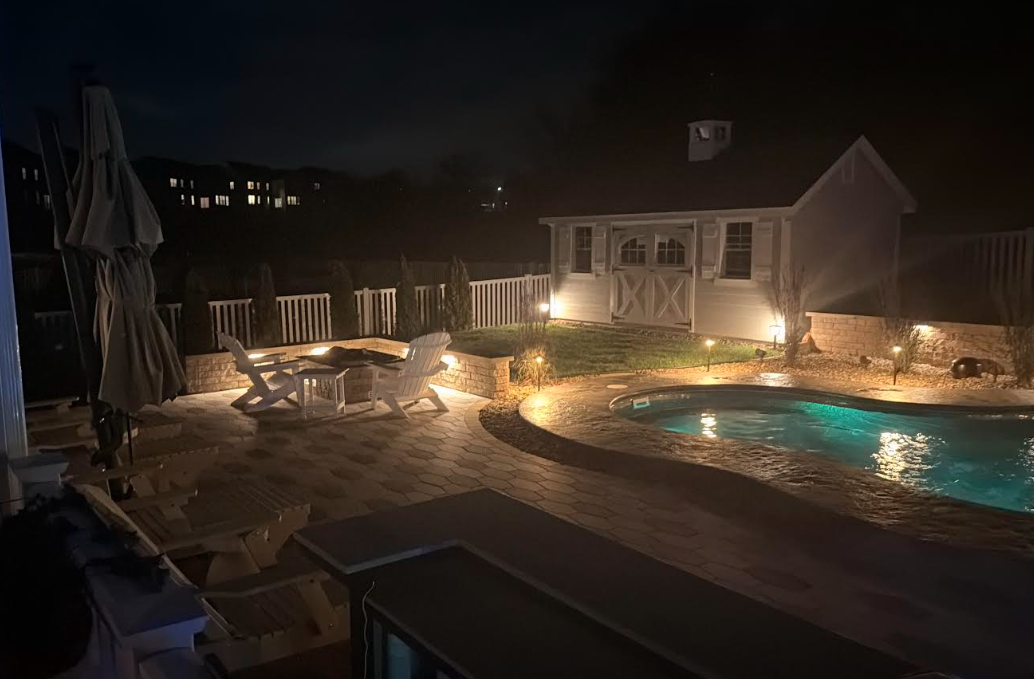 Night view of a backyard with a pool, patio, and illuminated small house.