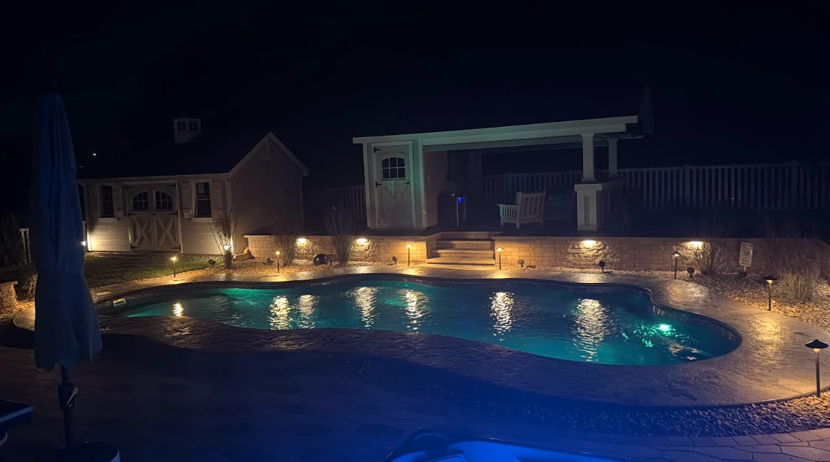 Nighttime view of a glowing pool with surrounding landscape lighting, a white pergola, and a small building in the background.