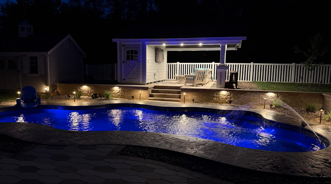 Backyard with a brick patio and a fire pit, overlooking a grassy lawn, white fence, and shed under a colorful sunset.