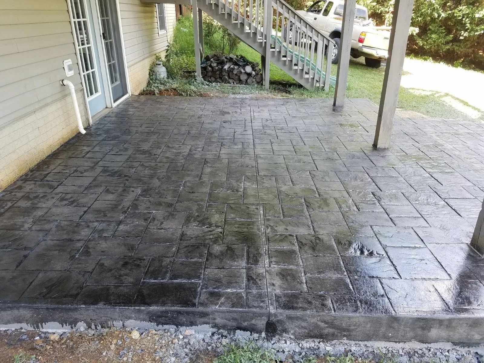 Gray, patterned concrete patio beneath a deck with a house on the left and grass on the right.