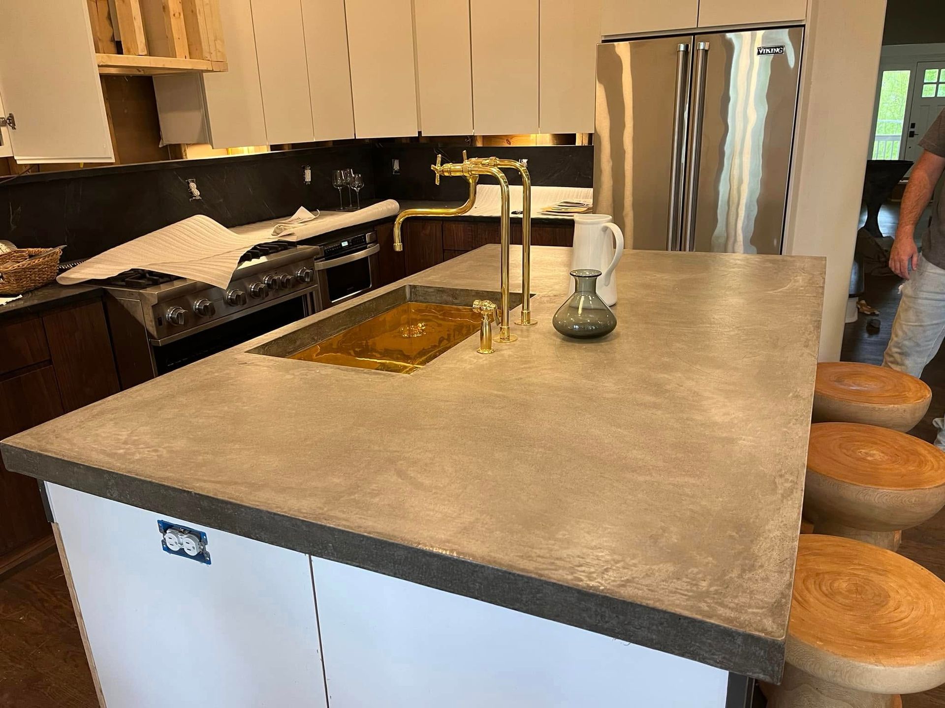 A kitchen island with a concrete countertop, gold faucet, and sink, surrounded by wooden stools, in a modern kitchen.
