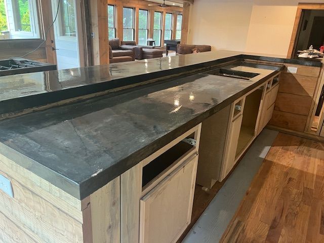 A kitchen island with a dark grey countertop and unfinished wood cabinetry under construction in a bright, open room.