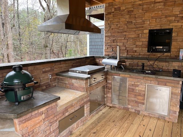 Outdoor kitchen with a green kamado grill, stainless steel appliances, and stone counters under a wooden roof.