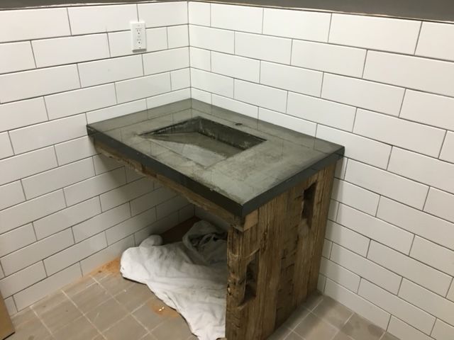 Concrete sink vanity with a rustic wooden base installed in a corner with white subway tile walls.