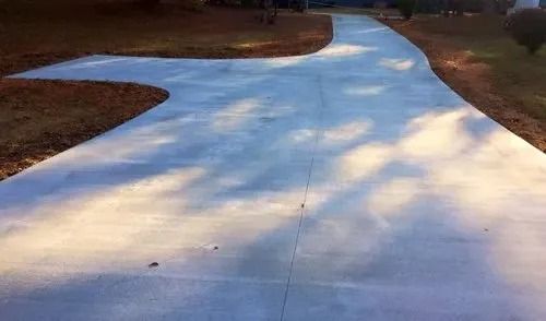 A newly poured light gray concrete driveway curves through a grassy lawn with dappled shadows from nearby trees.