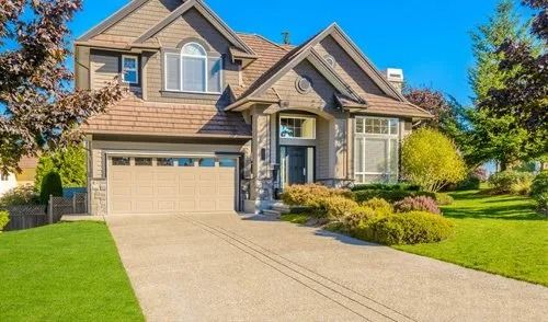 Two-story, beige stucco house with a shingled roof, attached garage, and paved driveway surrounded by green lawns.