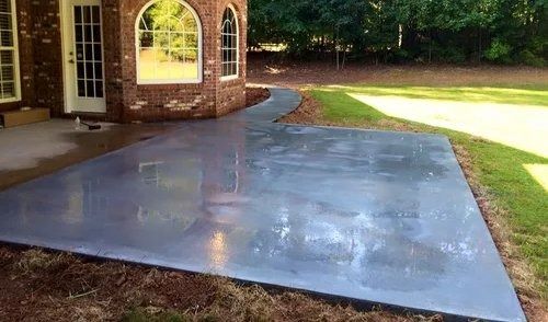A newly poured, smooth gray concrete patio extending from the back of a brick house into a green backyard.