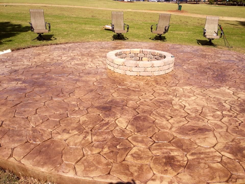 A circular stamped concrete patio with a stone fire pit in the center, featuring four chairs on a lawn in the background.