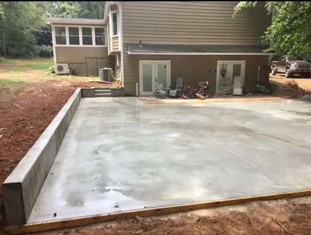 A newly poured concrete patio alongside a tan house with a retaining wall and surrounding red dirt.