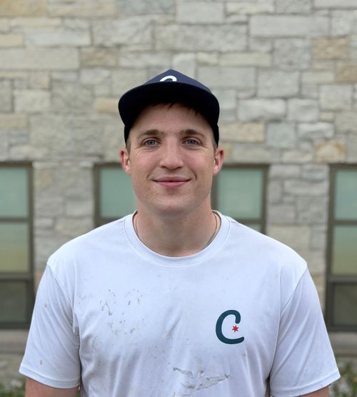 Smiling person in a white T-shirt and navy cap standing outdoors in front of a stone wall