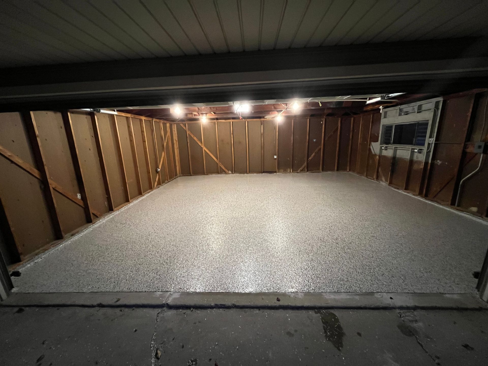 Empty garage with gray speckled epoxy floor and wooden walls, lit by overhead lights.