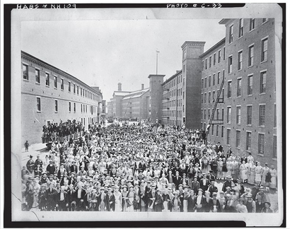 Large crowd of people outside brick mill buildings, possibly at an event or parade.