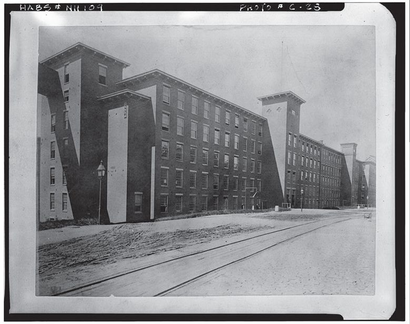 Large brick factory building with several stories and chimneys, with railroad tracks in front.