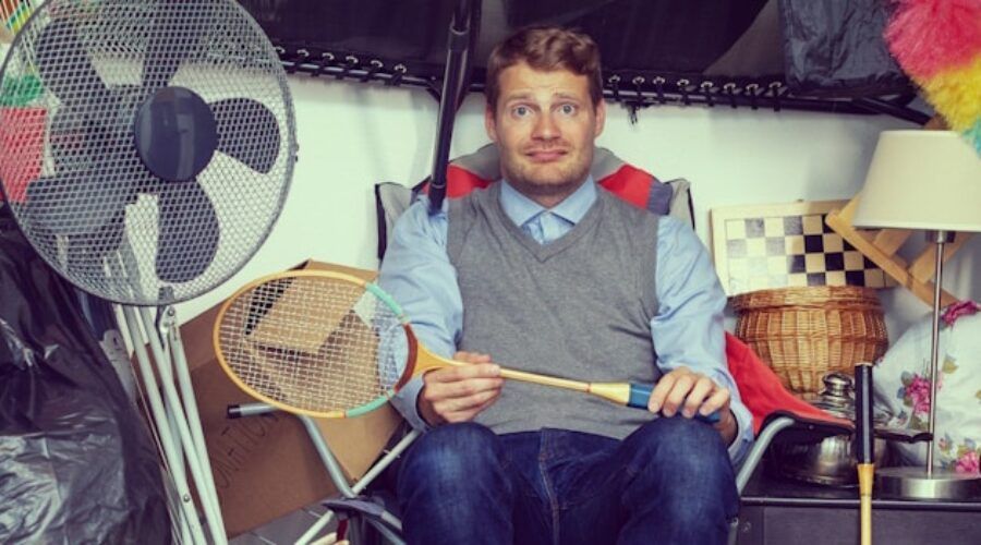 Man in a cluttered storage space, holding a badminton racket, looking surprised.