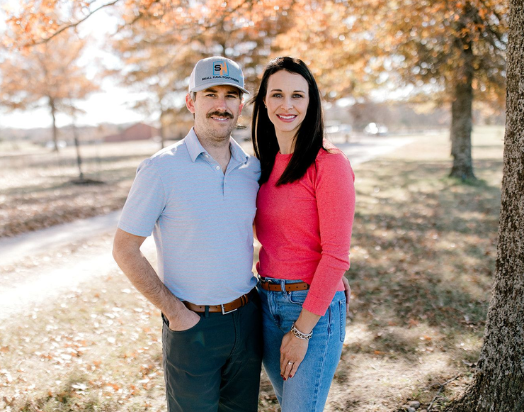 Couple standing outdoors; man in blue shirt and cap, woman in pink shirt, both smiling.