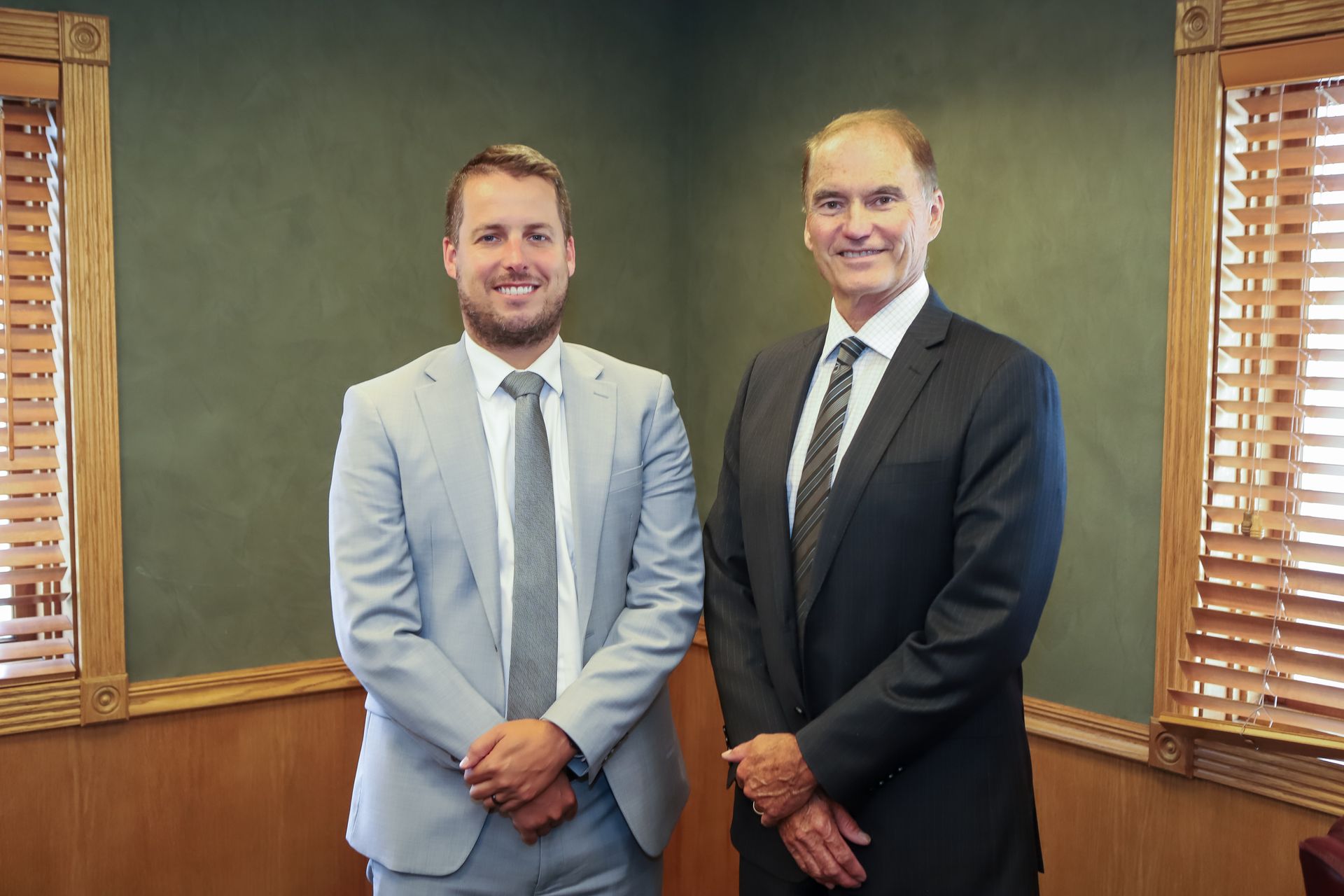 Two men in suits stand indoors, smiling slightly. Green wall, wood trim, and blinds inthe background.