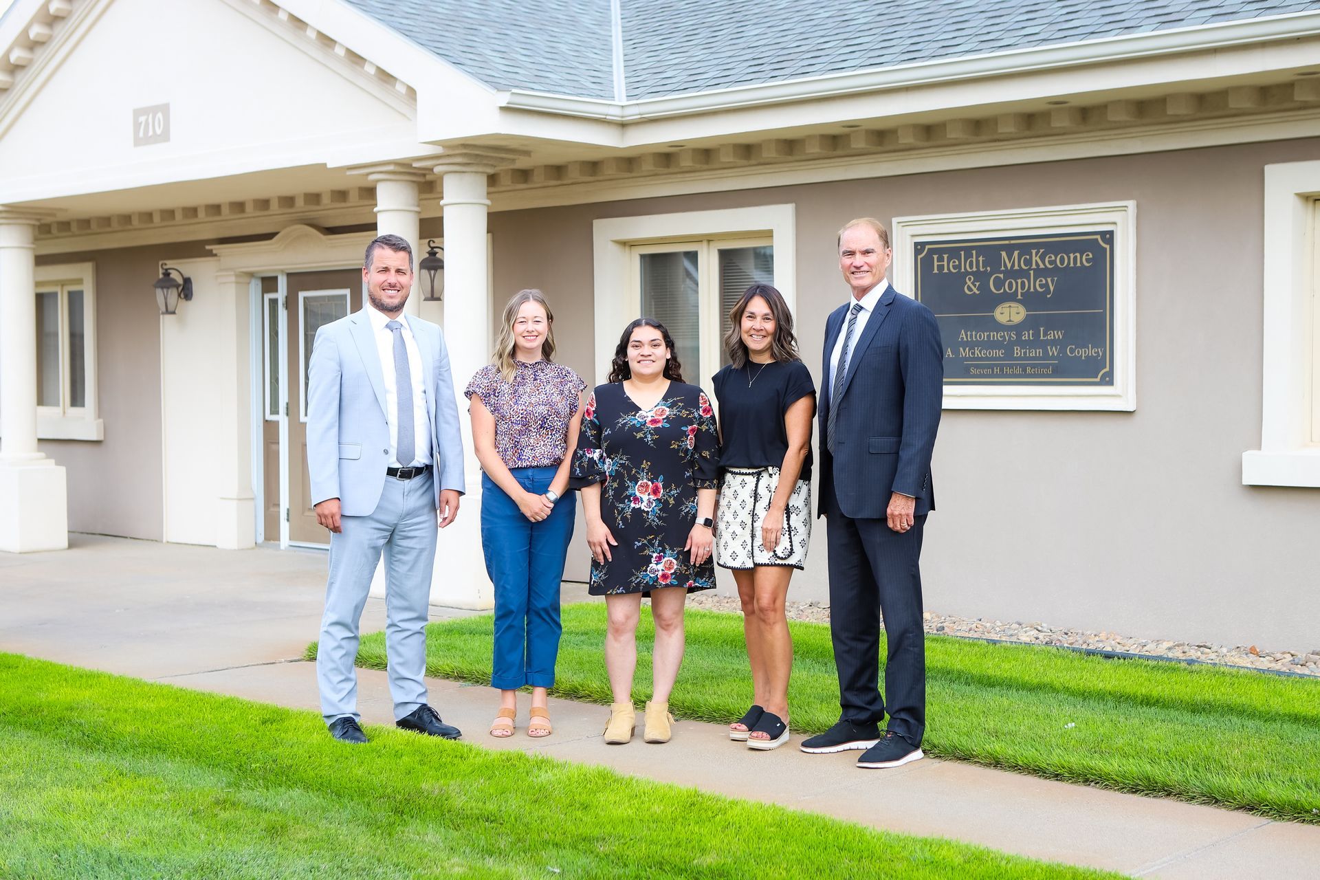 Five people stand in front of a building with a sign. The people are smiling.