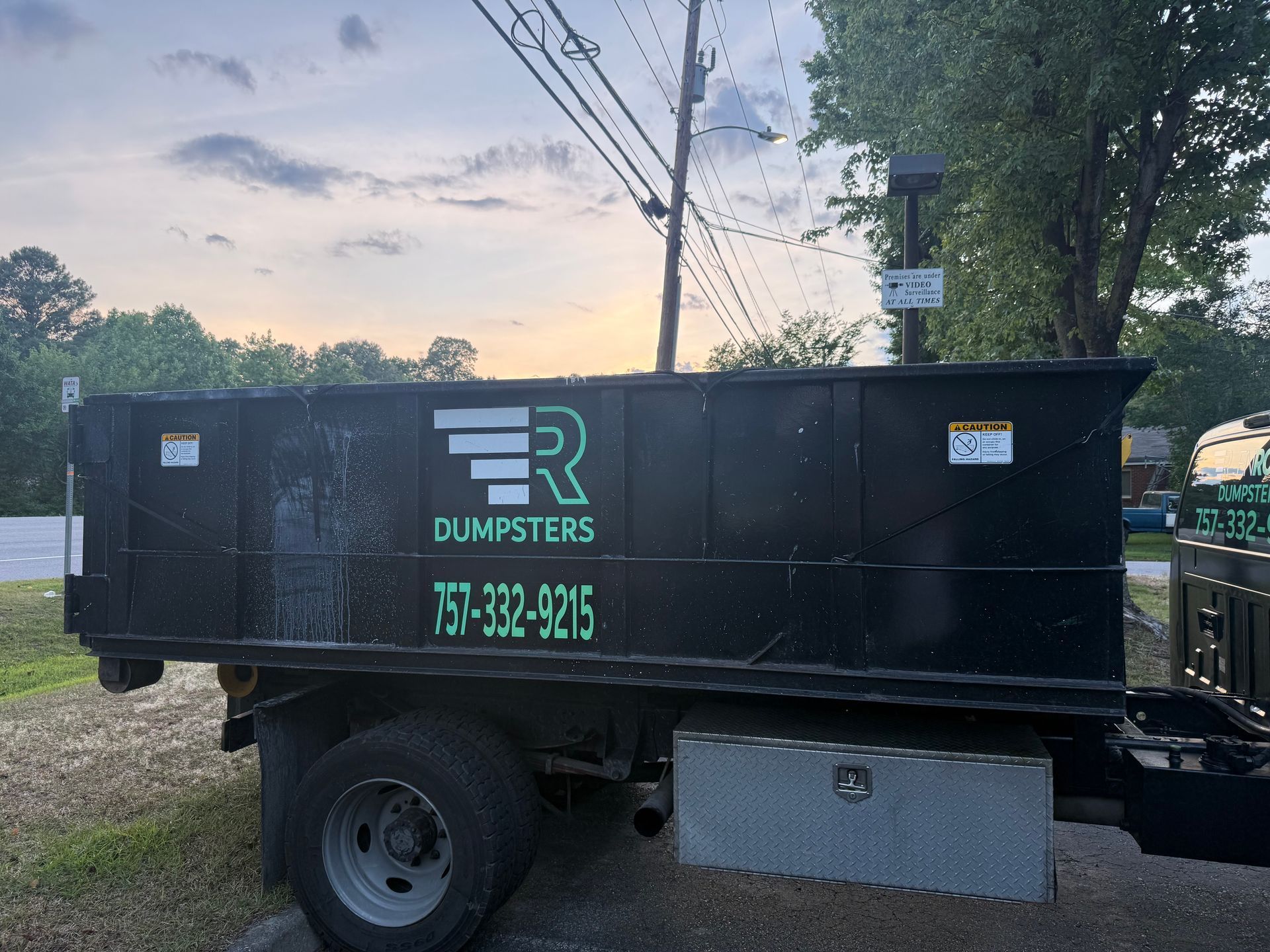 Black dumpster trailer parked outdoors near trees and power lines at dusk