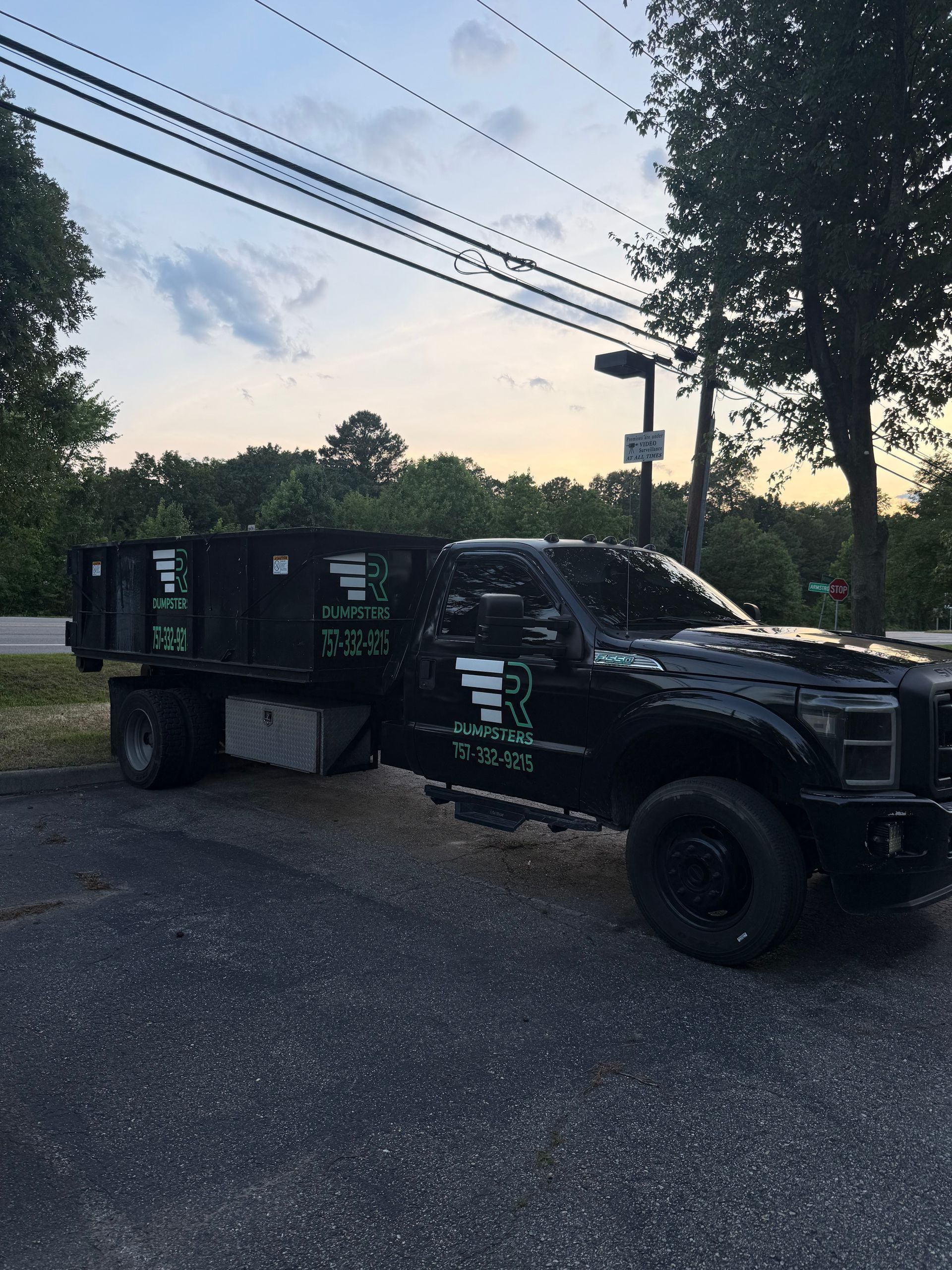 Black utility truck with dump bed parked on gravel at dusk, trees and power lines in the background