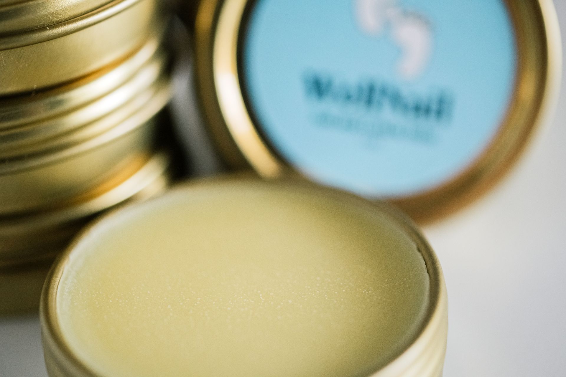 A close-up of a small, open tin of yellowish salve, with other tins stacked in the background, one with a blue label.