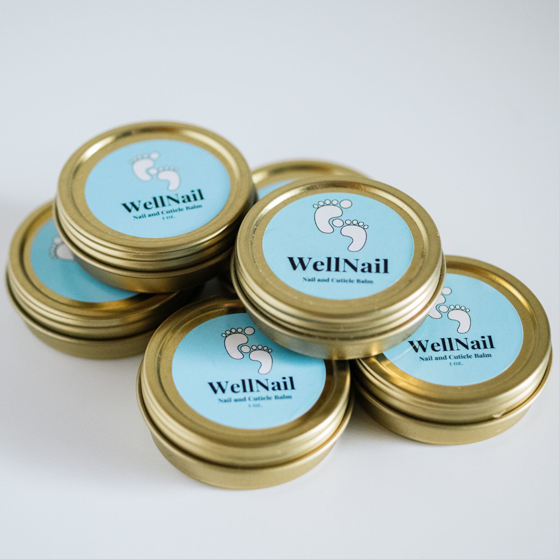 Six round gold tins of WellNail foot and nail balm, stacked on a white surface. Blue labels with baby feet.