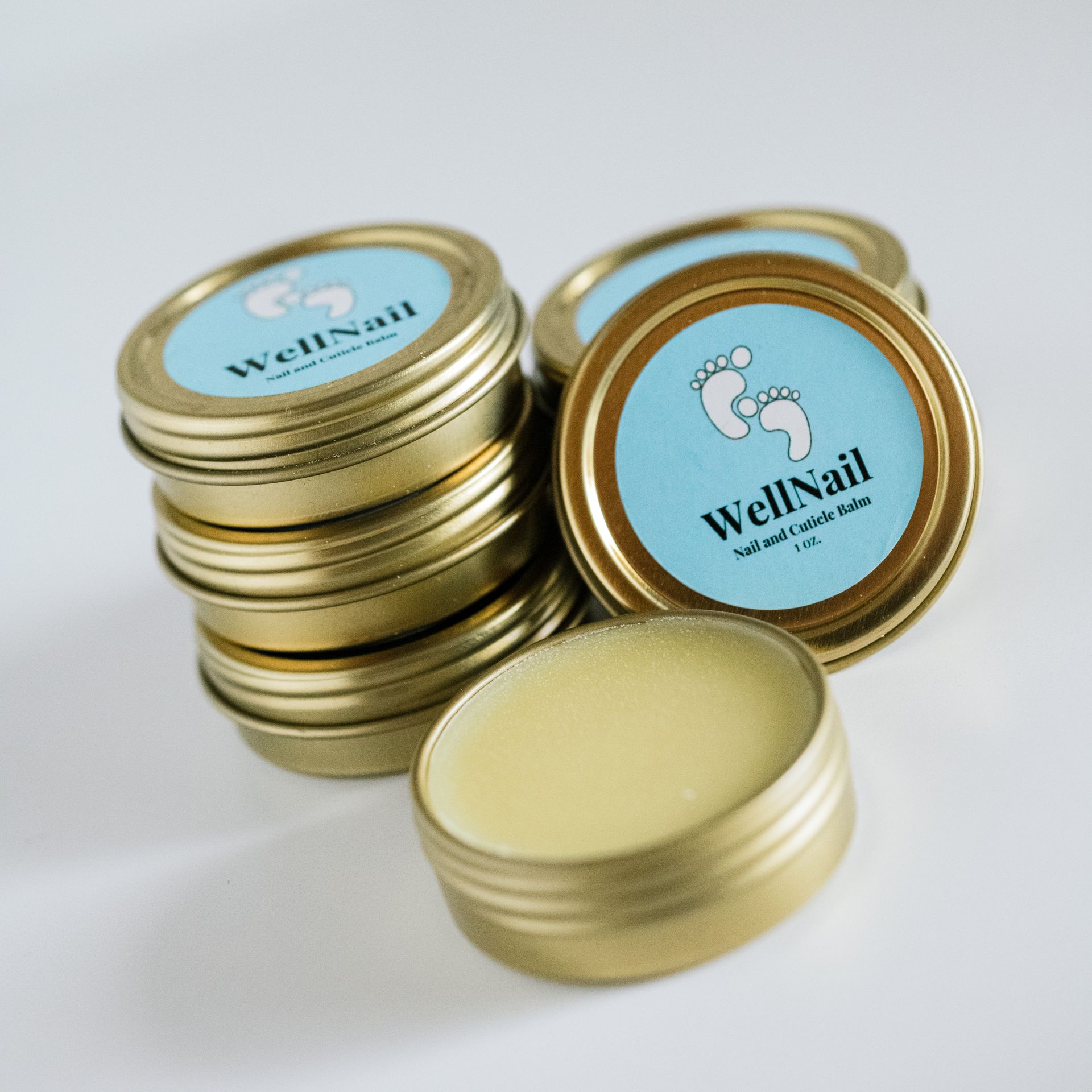 Small round gold tins of WellNail foot and cuticle butter stacked and opened, with a light blue label and white feet.