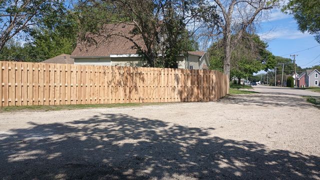 Wooden fence beside a gravel lot under trees on a sunny residential street
