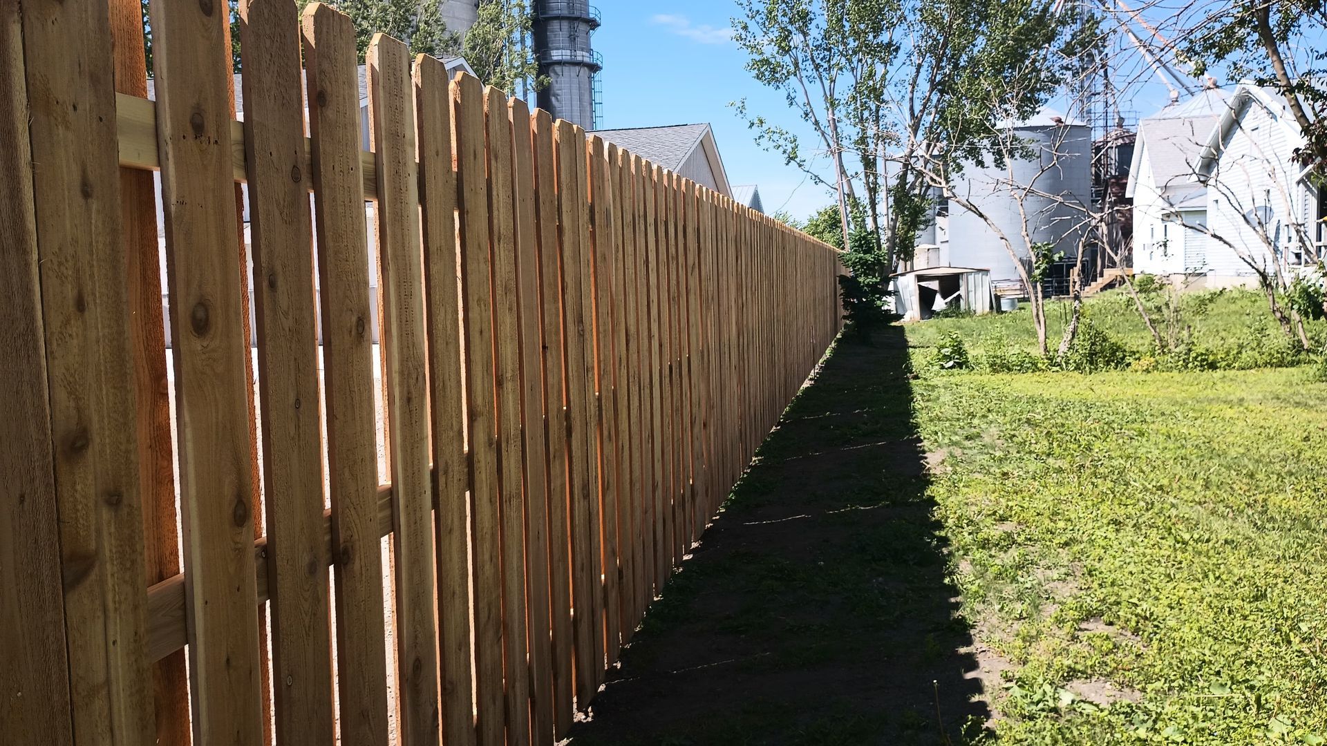 Wooden fence beside a sunlit grassy yard, with houses and trees in the background.