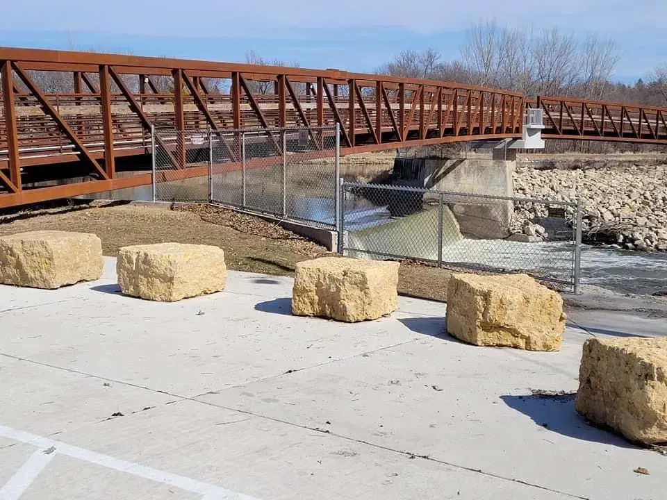 Rust-colored pedestrian bridge over a dry creek bed with large boulders in the foreground.