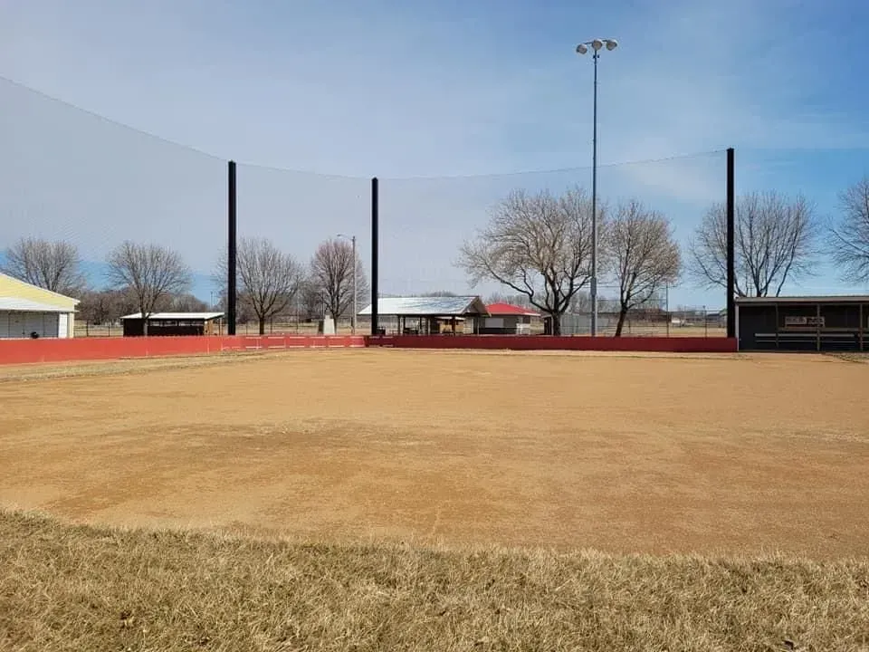 Empty baseball field with brown infield dirt, red outfield fence, and tall lights under a clear sky
