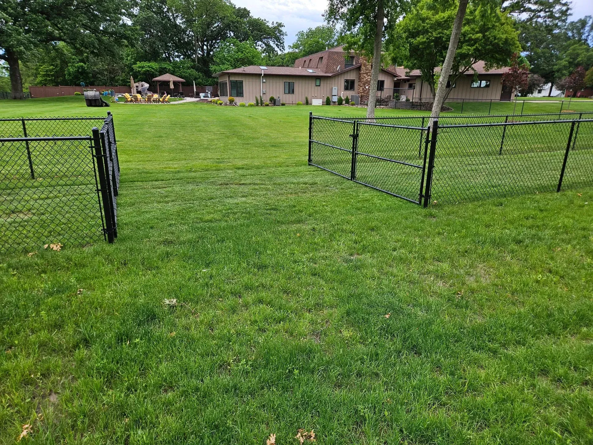 Grass field with two black chain-link fence panels in front of a house and trees.