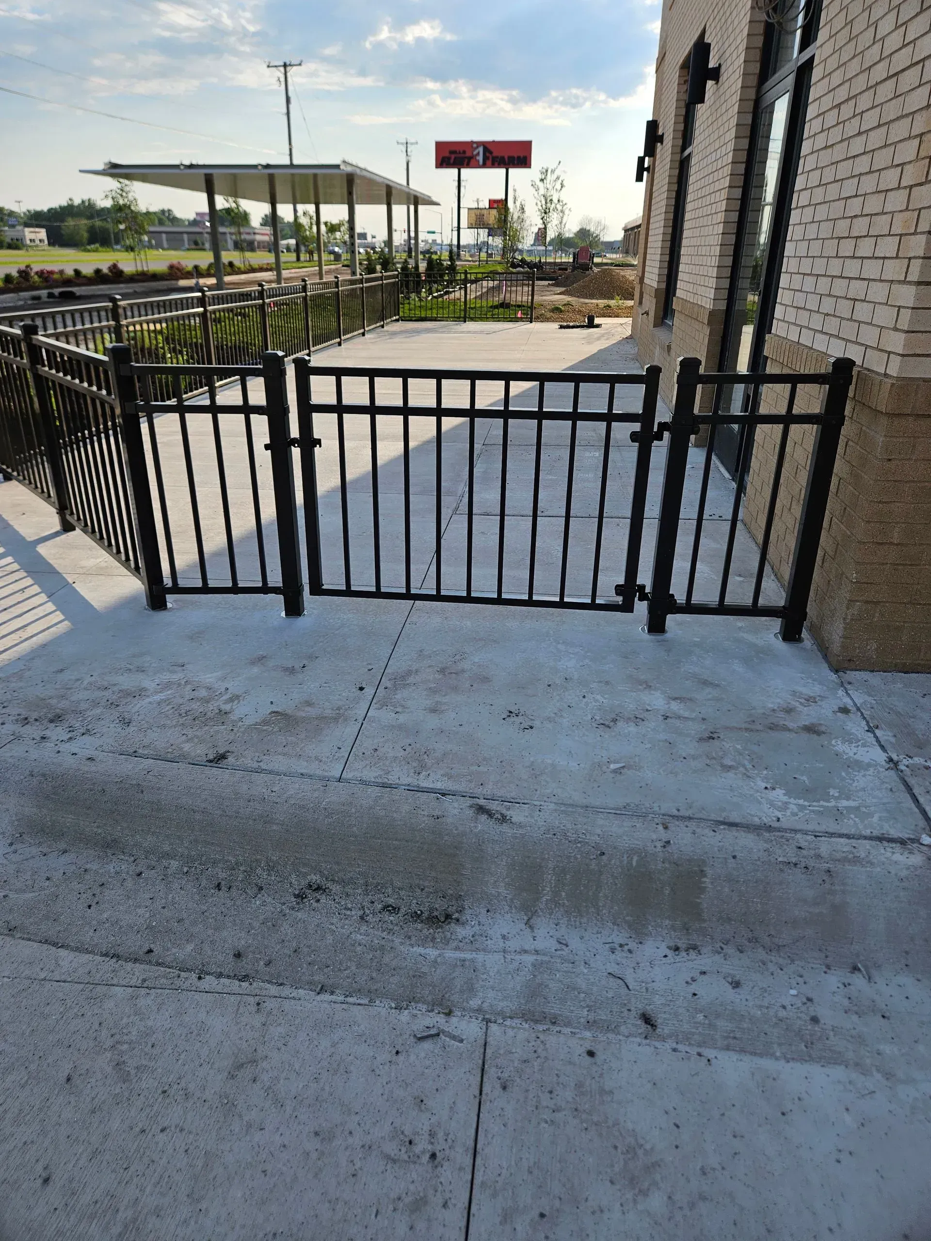 Black metal gate at a concrete walkway entrance beside a brick building, with a road and gas station in the background