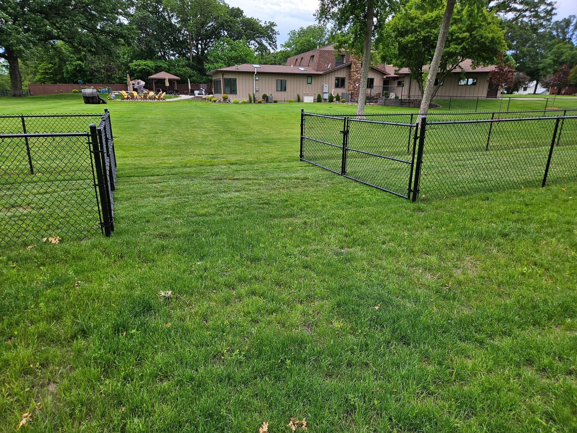 Open chain-link gate in a grassy yard with a house and trees in the background