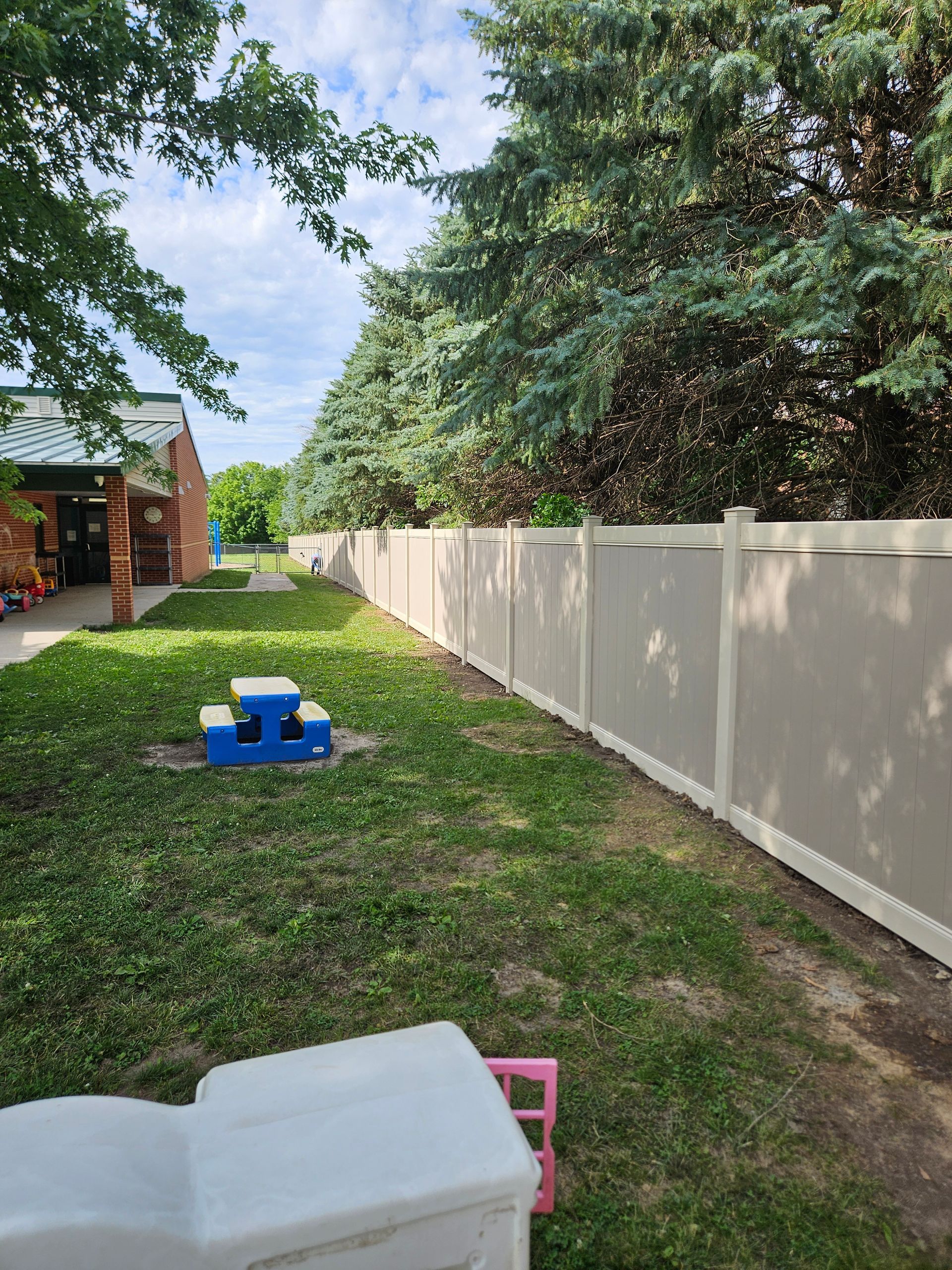 Backyard with a white fence, green lawn, trees, and children’s play equipment near a patio