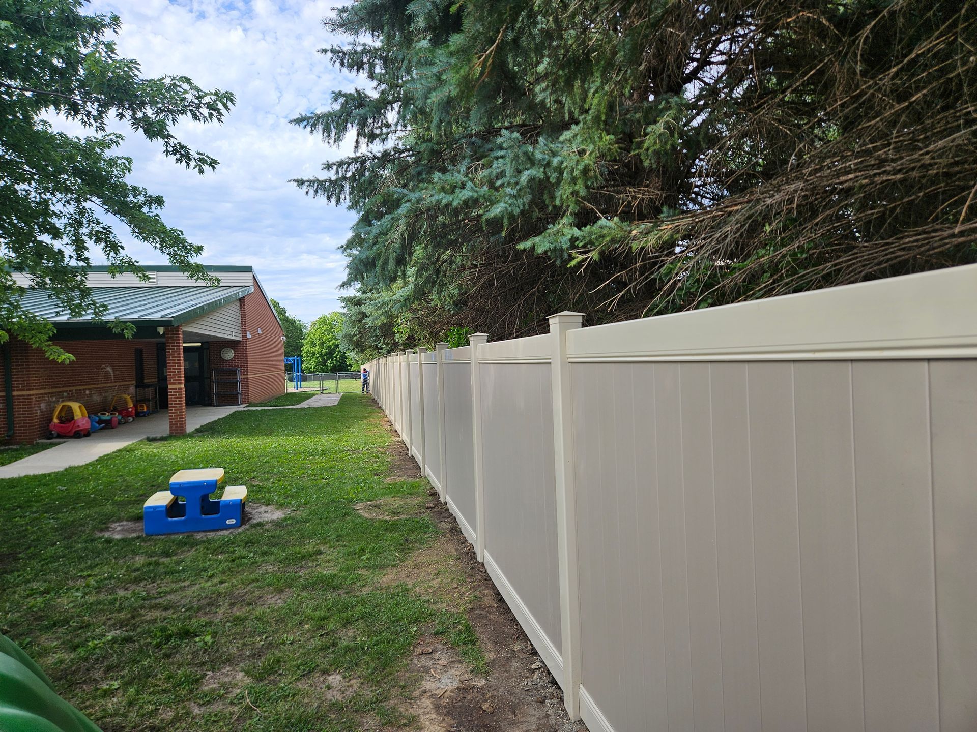Backyard with a long beige fence, grassy lawn, brick house, and blue toy near a tree-lined path