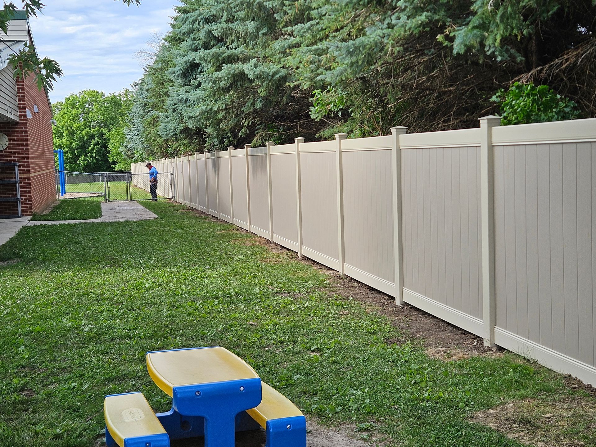 Grassy alley beside beige privacy fence and brick building, with blue-and-yellow picnic table in foreground