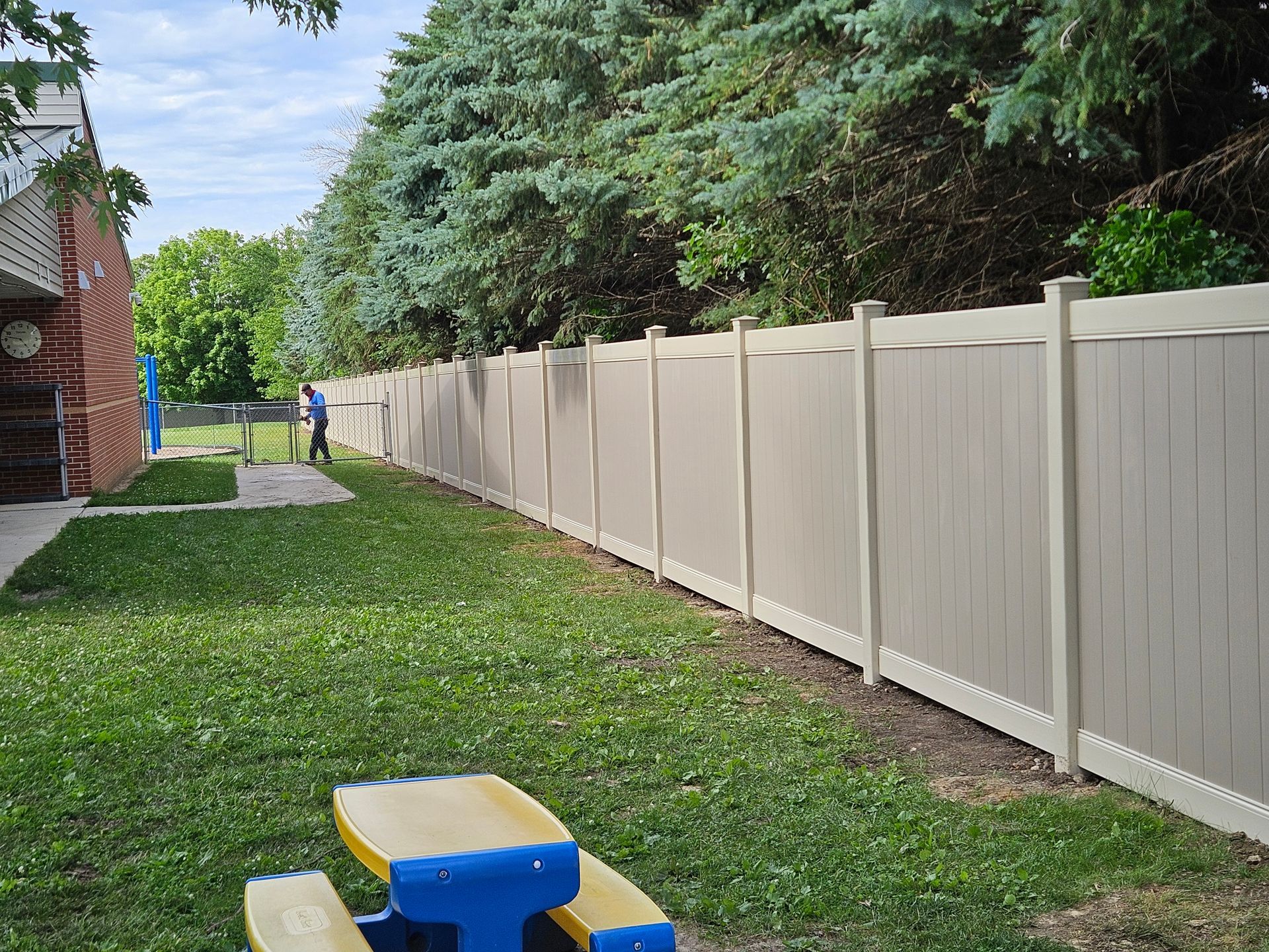 Backyard with green lawn, long white fence, and blue-yellow plastic play table in foreground.