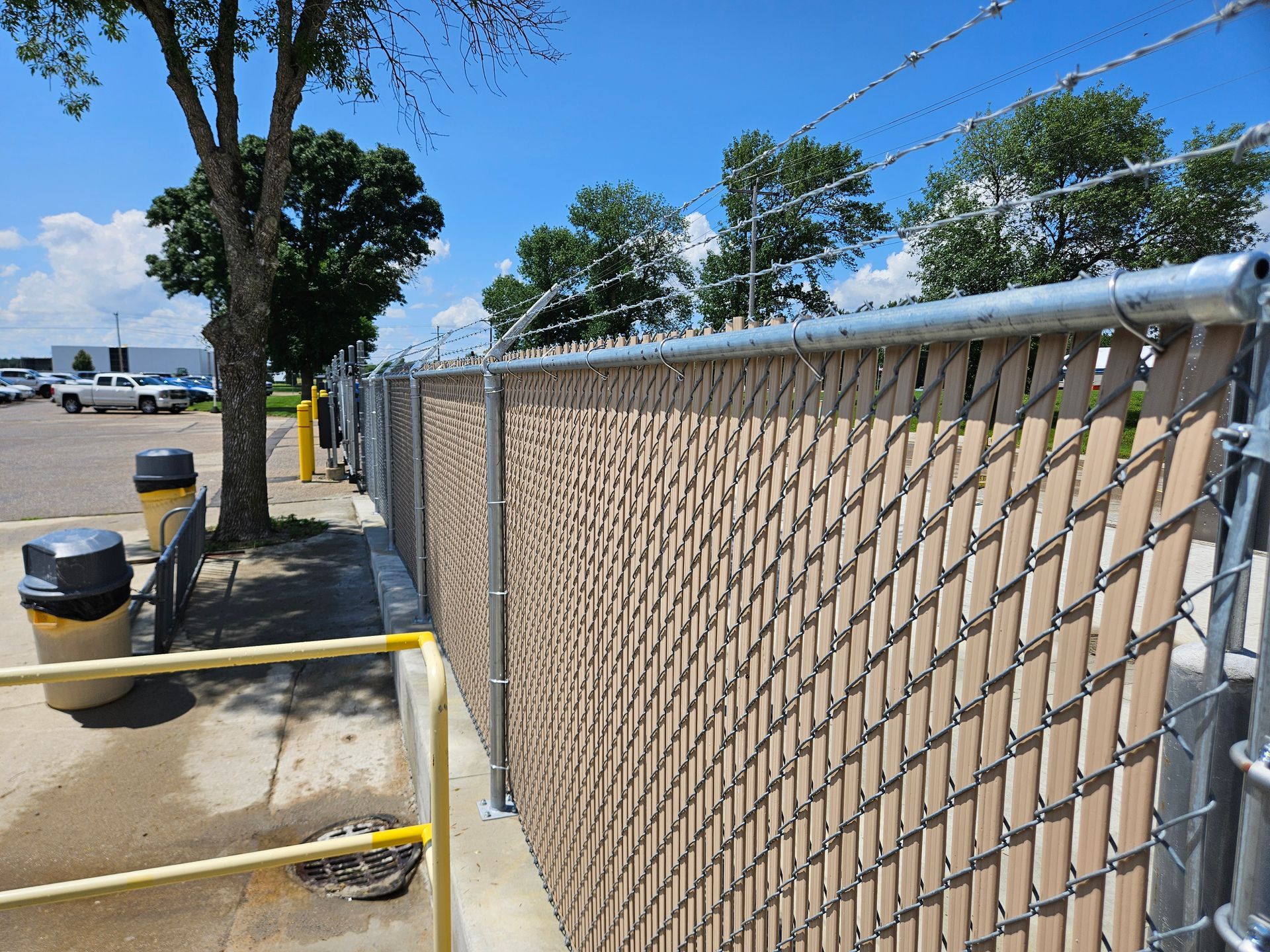Chain-link fence along a sunny sidewalk beside a street and parked vehicles