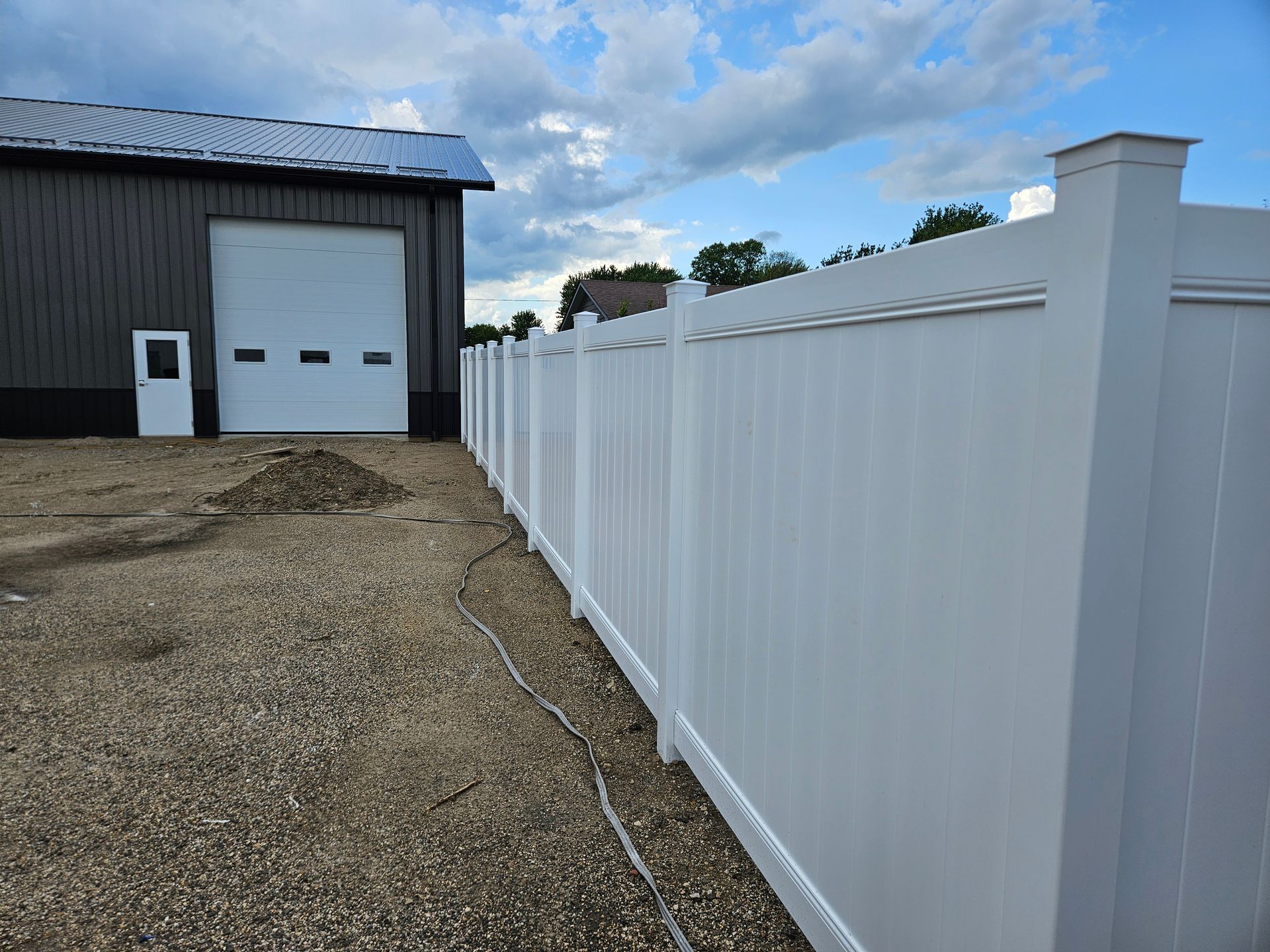 White vinyl fence along a gravel driveway beside a warehouse with a garage door under a cloudy sky