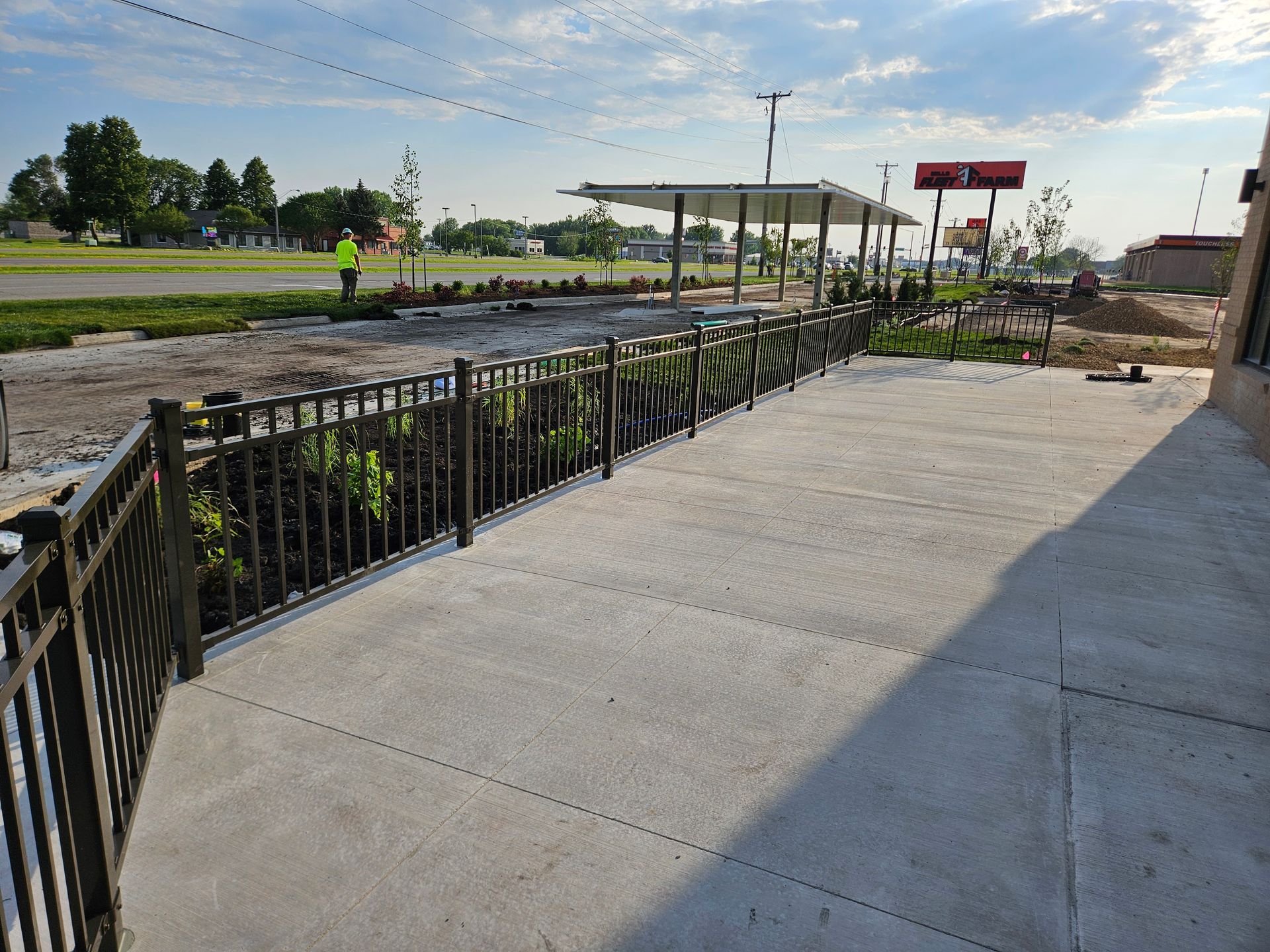 Sidewalk beside a fenced construction site with concrete pavement, plants, and a small shelter in the distance