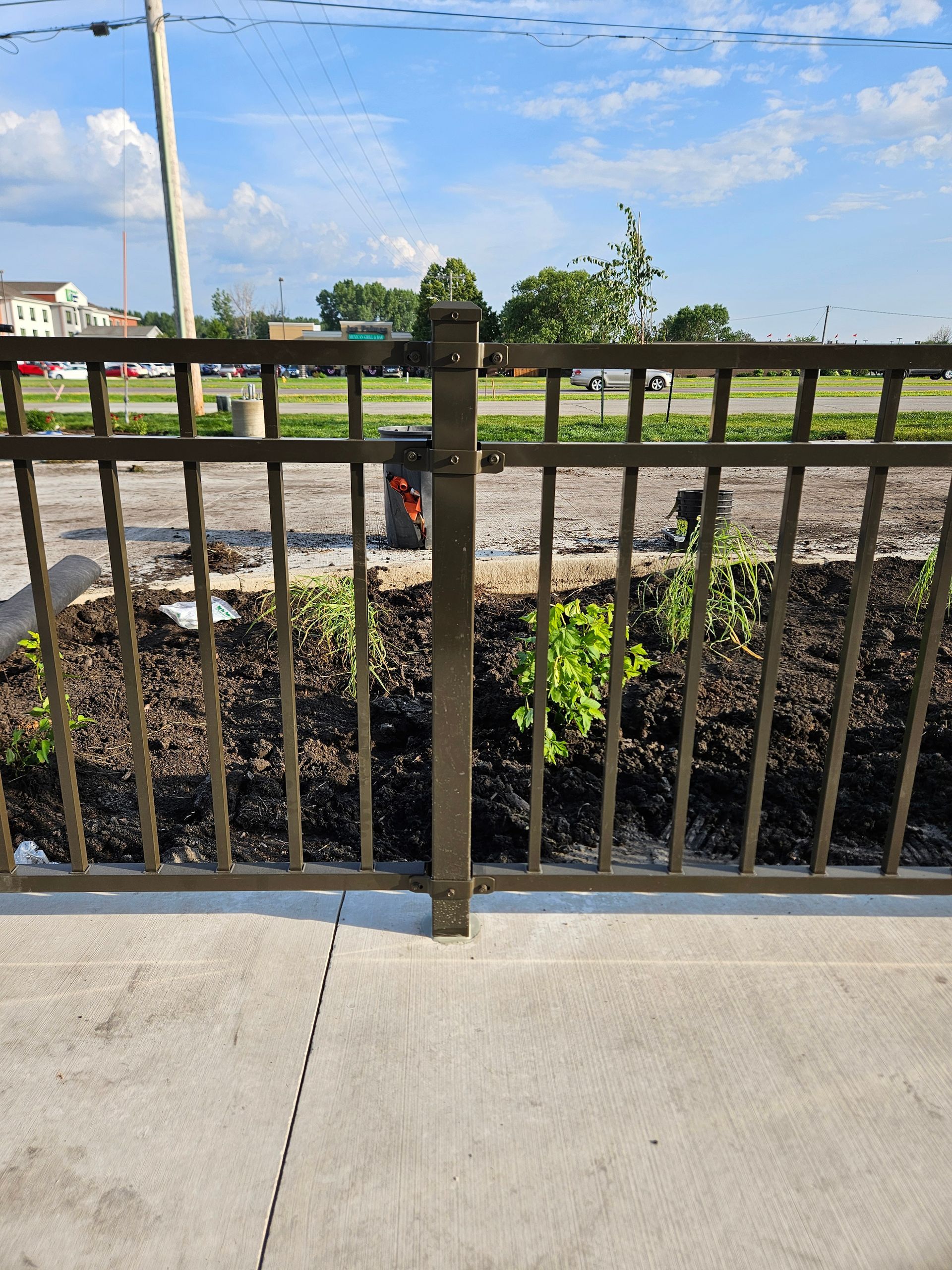 Black metal fence along a concrete sidewalk with a road, trees, and blue sky in the background