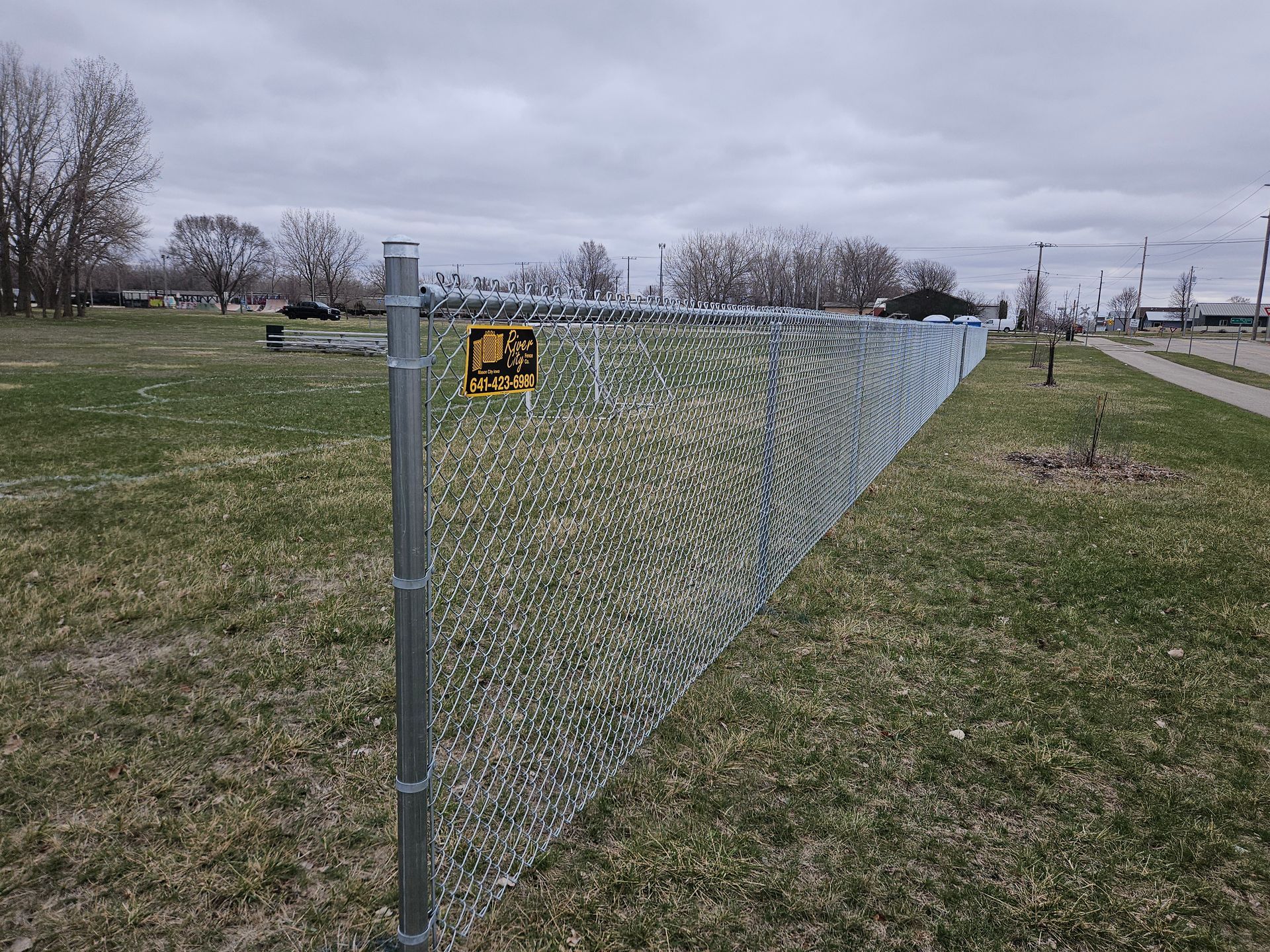 Chain-link fence along a grassy field under an overcast sky, with a yellow sign in the distance.