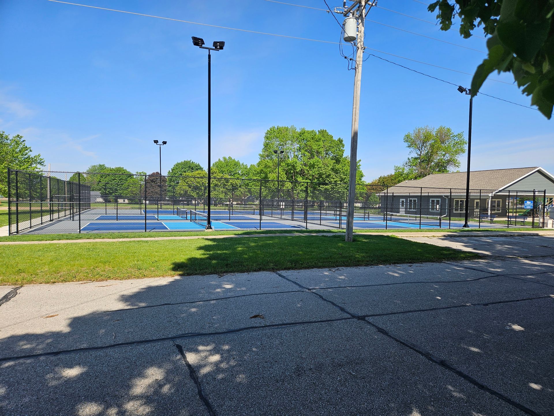 Outdoor tennis courts with black fencing, blue playing surface, and trees under a clear sky.