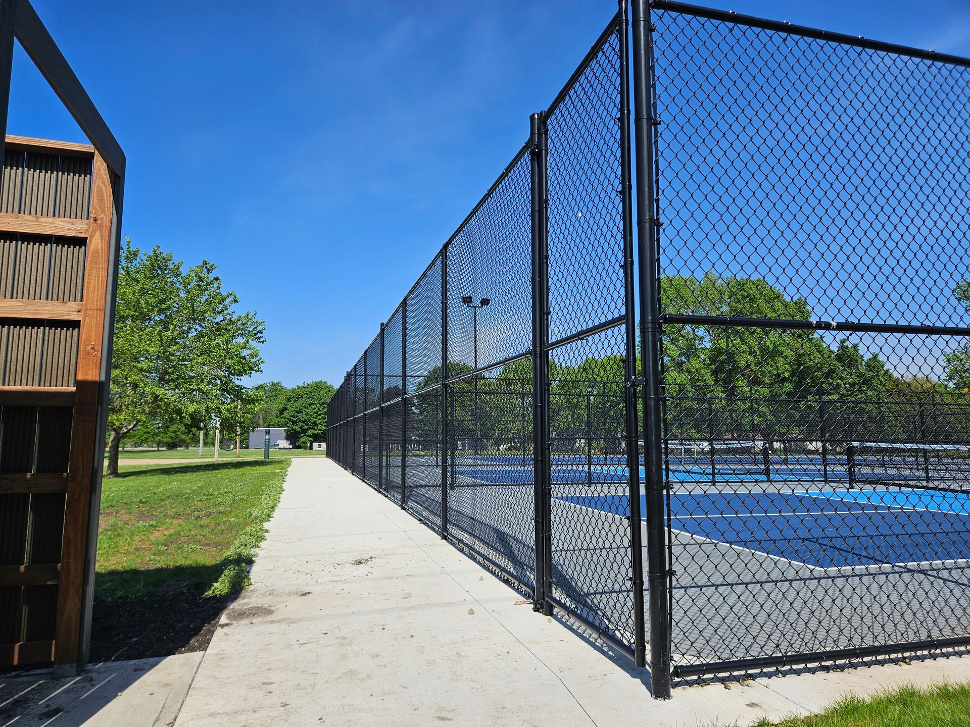 Fenced blue tennis courts beside a sidewalk and trees under a clear blue sky
