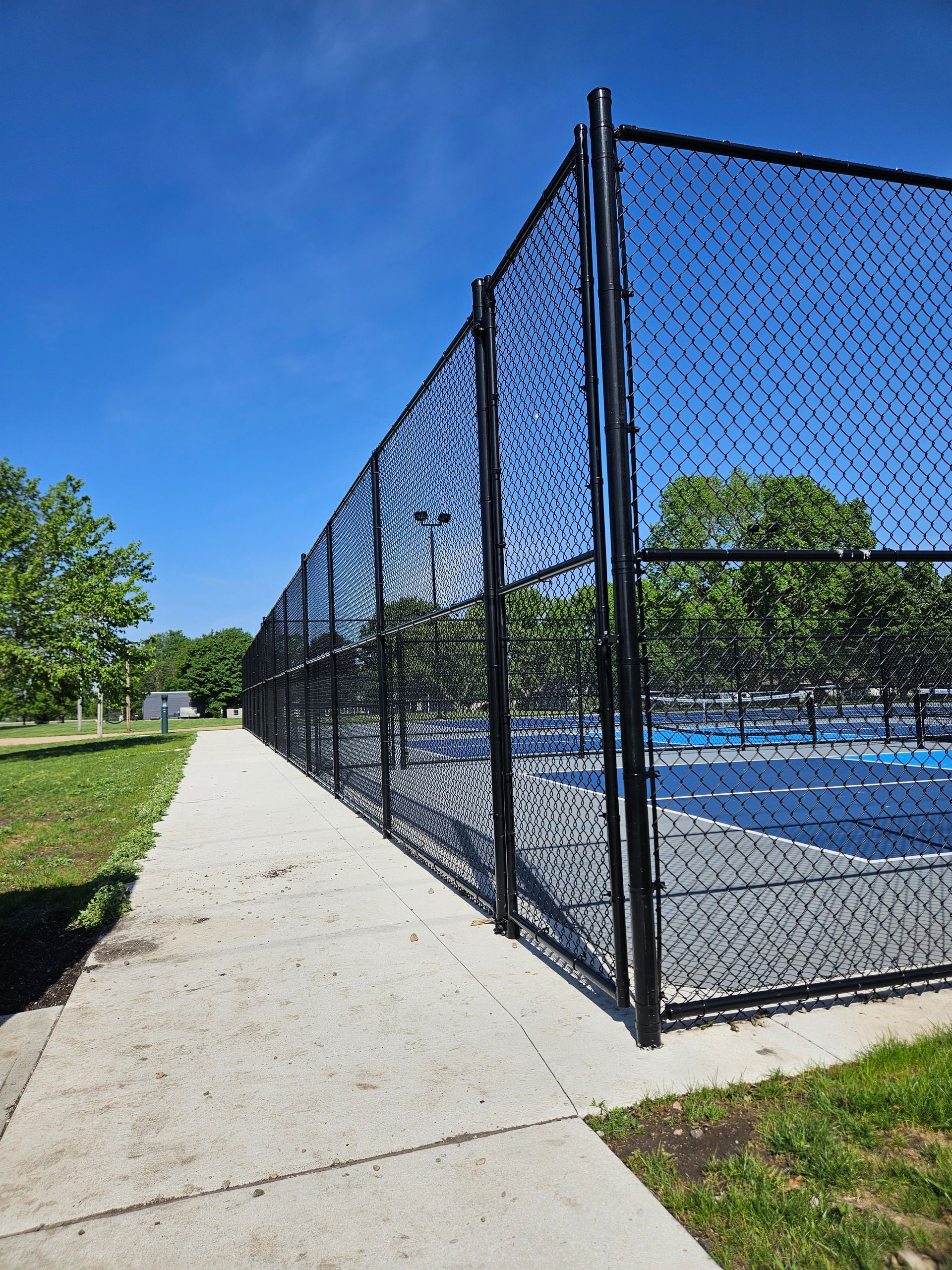 Chain-link fence beside a blue tennis court along a concrete path under a clear sky