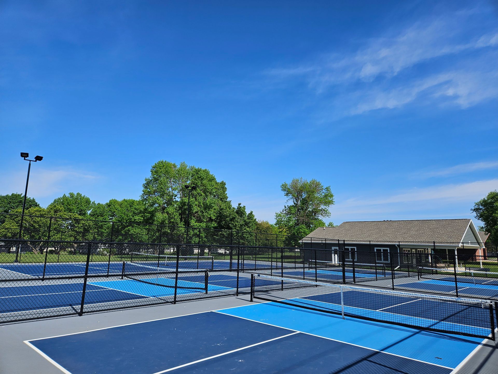 Blue tennis courts with black fencing and trees under a clear blue sky