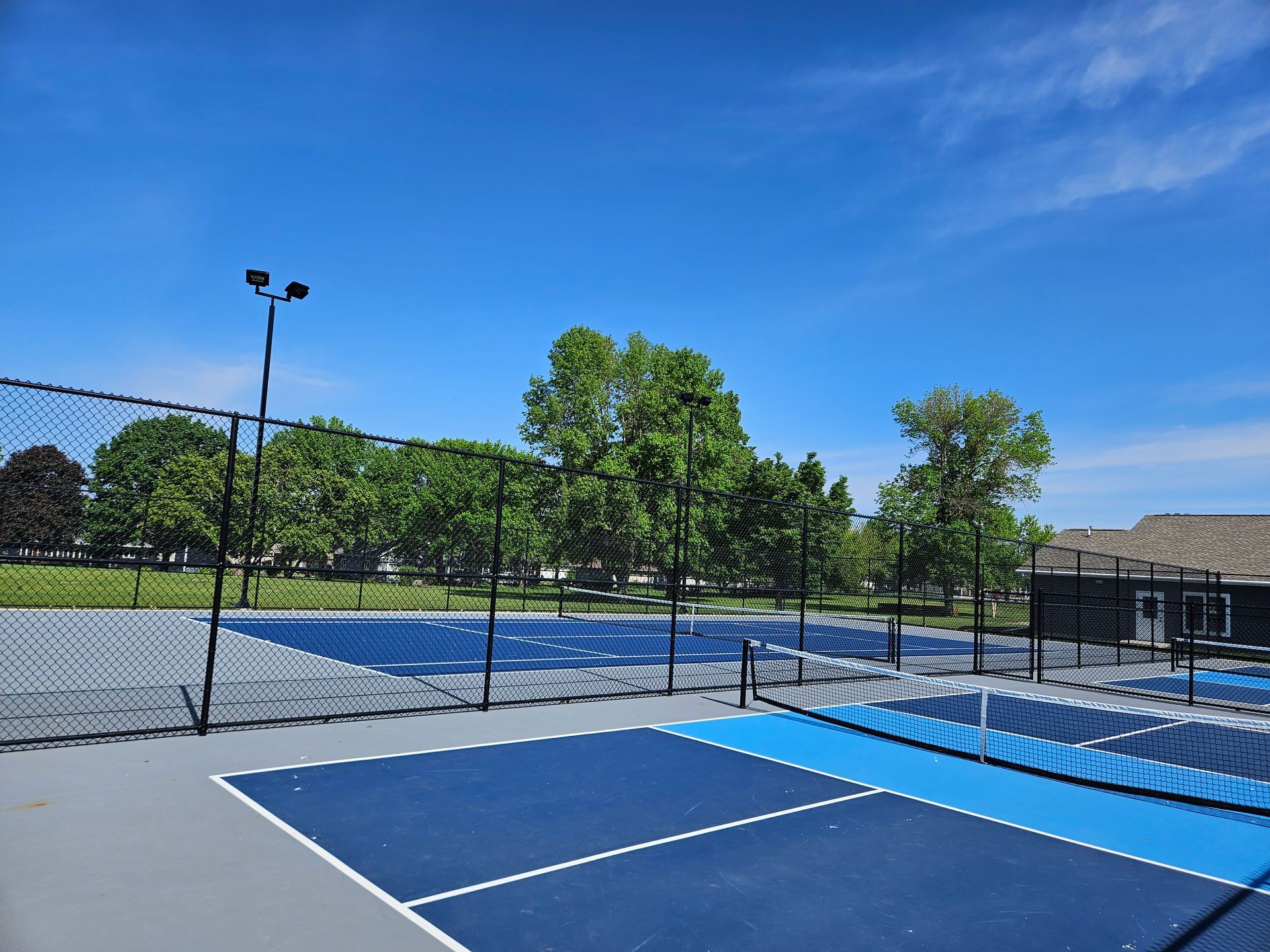 Empty blue tennis courts beside a chain-link fence under a clear sky, with trees and lamp posts nearby