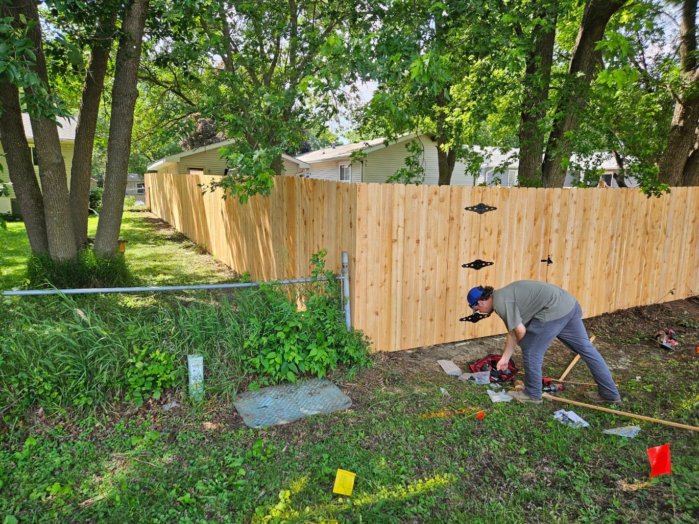 Person digging in a grassy backyard beside a wooden fence, with tools and bright flags visible.